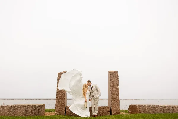 A bride and groom kissing in front of stone pillars near a body of water.