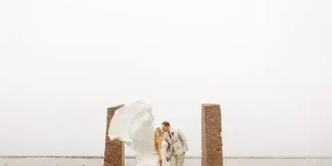 A bride and groom kissing in front of stone pillars near a body of water.