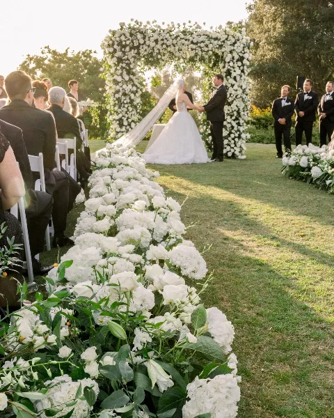 Outdoor wedding ceremony with a white floral arch and aisle runner of white roses and hydrangeas.