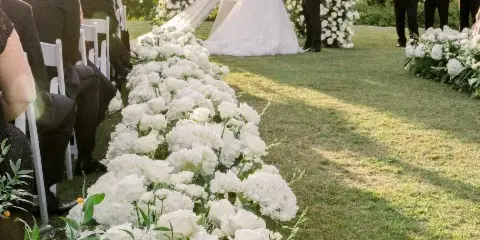 Outdoor wedding ceremony with a white floral arch and aisle runner of white roses and hydrangeas.