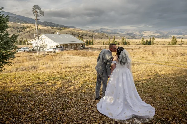 A bride and groom kissing in a field in front of a white barn with mountains in the background.