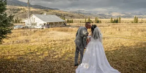 A bride and groom kissing in a field in front of a white barn with mountains in the background.