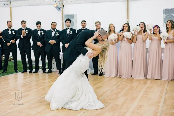A bride and groom share a dip during their first dance while the wedding party watches and claps in a white tent.