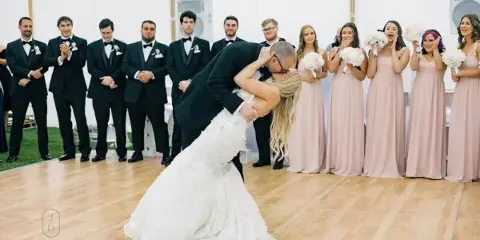 A bride and groom share a dip during their first dance while the wedding party watches and claps in a white tent.