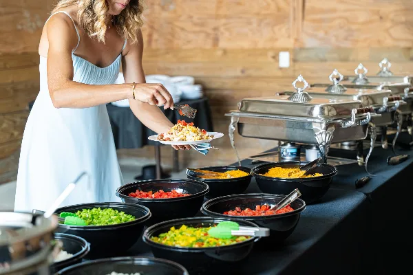 A woman in a light blue dress serves herself from a buffet line with chafing dishes and bowls of prepared food at a wedding reception.