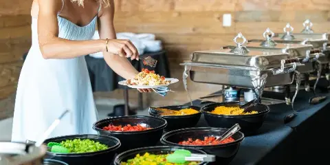 A woman in a light blue dress serves herself from a buffet line with chafing dishes and bowls of prepared food at a wedding reception.
