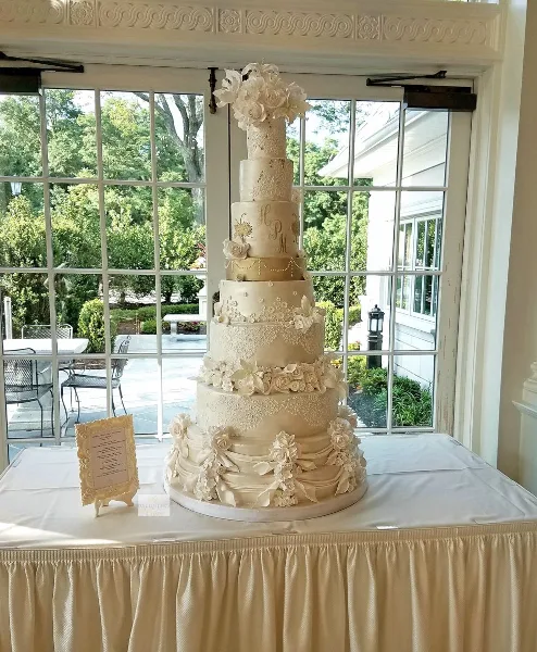 A tall, multi-tiered white wedding cake decorated with sugar flowers and lace piping, displayed on a table in front of a window.