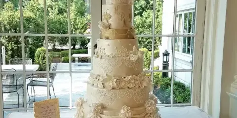 A tall, multi-tiered white wedding cake decorated with sugar flowers and lace piping, displayed on a table in front of a window.
