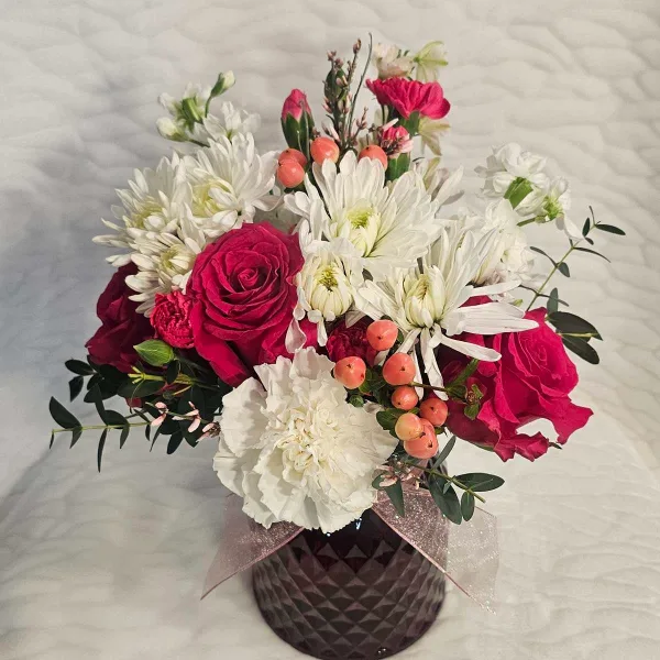 A floral arrangement featuring deep red roses, white chrysanthemums, and orange berries in a dark faceted vase.