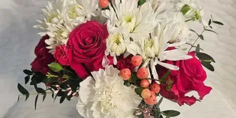 A floral arrangement featuring deep red roses, white chrysanthemums, and orange berries in a dark faceted vase.
