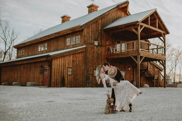 A bride and groom kissing in front of a large rustic wooden barn wedding venue.