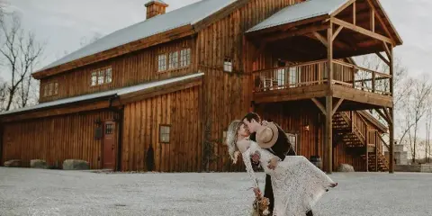 A bride and groom kissing in front of a large rustic wooden barn wedding venue.