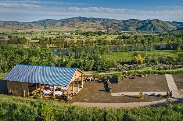 Aerial view of a rustic wooden barn with a metal roof situated on a ridge overlooking a river valley and mountains.