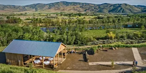 Aerial view of a rustic wooden barn with a metal roof situated on a ridge overlooking a river valley and mountains.