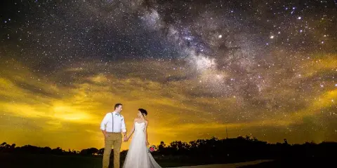 A bride and groom standing hand-in-hand under a starry night sky with the Milky Way visible.