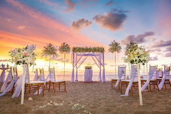 A beach wedding ceremony setup featuring a floral arch, chairs, and floral stands against a sunset ocean backdrop.