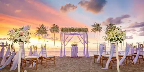 A beach wedding ceremony setup featuring a floral arch, chairs, and floral stands against a sunset ocean backdrop.
