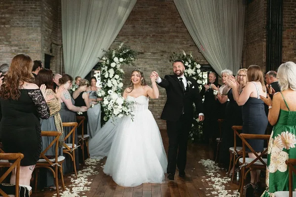 A bride and groom walking down the aisle during a wedding reception in a venue with exposed brick walls and floral arches.
