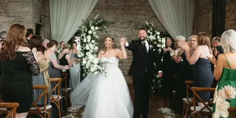A bride and groom walking down the aisle during a wedding reception in a venue with exposed brick walls and floral arches.