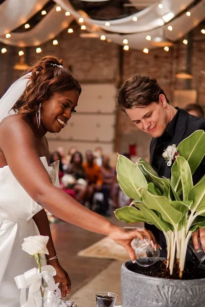 A bride and groom smiling while participating in a wedding ritual involving a potted plant, with guests seated in the background.