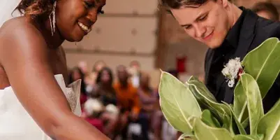 A bride and groom smiling while participating in a wedding ritual involving a potted plant, with guests seated in the background.