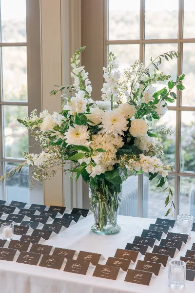A tall white floral centerpiece in a glass vase sitting on a table with place cards.