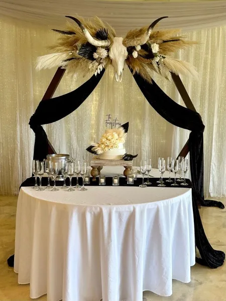 A wedding event setup featuring a sweetheart table with a white tablecloth, champagne flutes, and a decorative backdrop with a skull, horns, and dried florals.