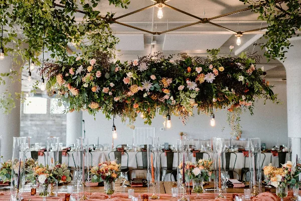 A large, lush floral installation with hanging greenery and mixed flowers suspended above a long event table.
