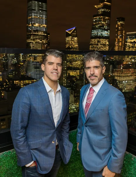 Two men in blue suits posing for a professional portrait against a city skyline background.