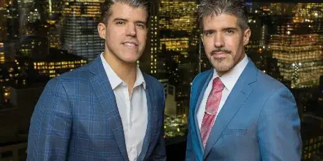 Two men in blue suits posing for a professional portrait against a city skyline background.