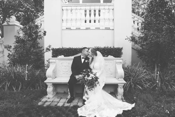 A black and white photograph of a bride and groom kissing while seated on an ornate stone bench in a garden setting.