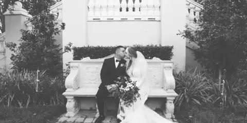 A black and white photograph of a bride and groom kissing while seated on an ornate stone bench in a garden setting.
