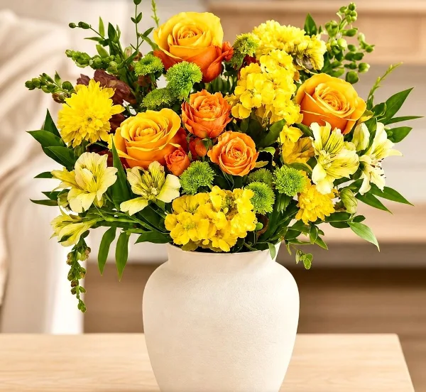 A vibrant floral arrangement featuring yellow and orange roses, alstroemeria, and greenery in a white ceramic vase.