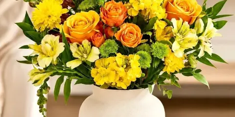 A vibrant floral arrangement featuring yellow and orange roses, alstroemeria, and greenery in a white ceramic vase.
