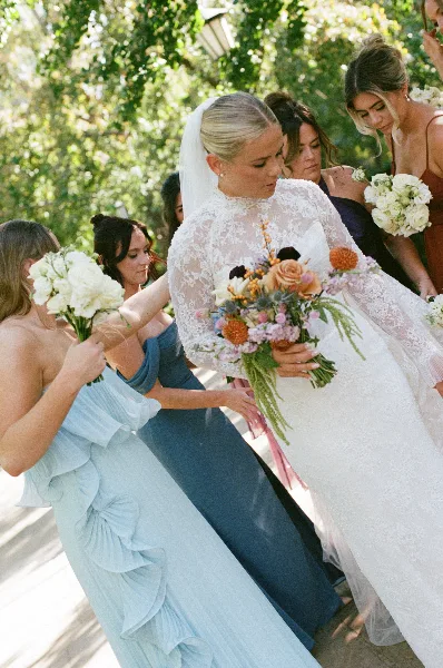 A bride in a white lace gown holding a bouquet, surrounded by bridesmaids in blue dresses outdoors.