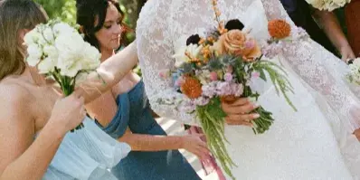 A bride in a white lace gown holding a bouquet, surrounded by bridesmaids in blue dresses outdoors.