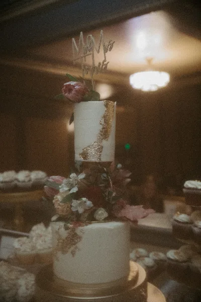 A two-tier white wedding cake decorated with gold leaf and fresh flowers, topped with a 'Mr & Mrs' topper, displayed on a stand with cupcakes in the background.