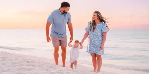 A family of three walking on a sandy beach at sunset.