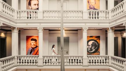 Interior view of a luxury hotel lobby featuring white classical columns, a balcony, and large portrait art on the walls.
