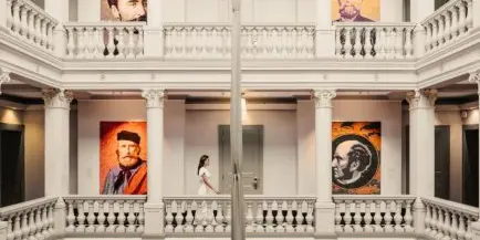 Interior view of a luxury hotel lobby featuring white classical columns, a balcony, and large portrait art on the walls.