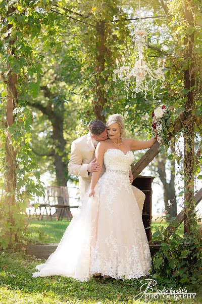 A bride in a white lace gown and a groom in a beige suit embracing under a wooden archway decorated with greenery and a crystal chandelier.