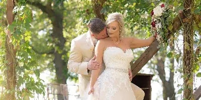 A bride in a white lace gown and a groom in a beige suit embracing under a wooden archway decorated with greenery and a crystal chandelier.