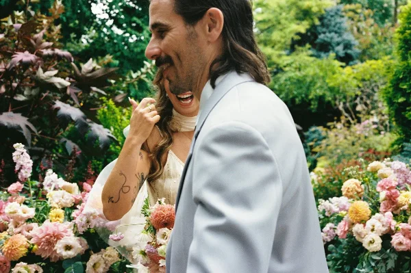 A candid film photograph of a bride and groom laughing together in a lush garden filled with colorful flowers.