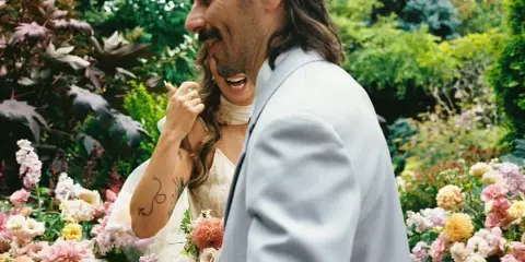 A candid film photograph of a bride and groom laughing together in a lush garden filled with colorful flowers.