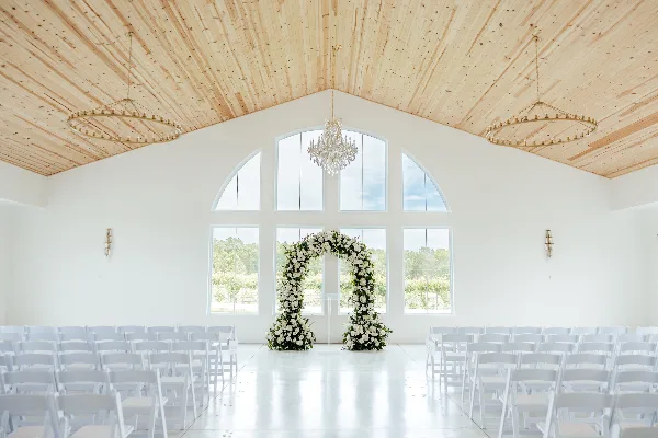 Interior of a white wedding venue with a wooden ceiling, large arched windows, a floral arch, and rows of white chairs.