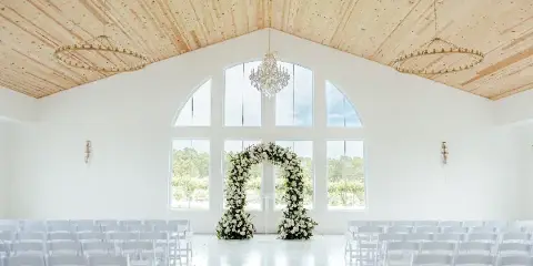Interior of a white wedding venue with a wooden ceiling, large arched windows, a floral arch, and rows of white chairs.