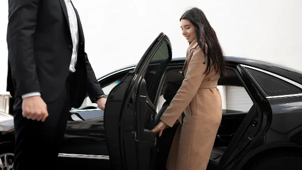 A professional chauffeur in a black suit opens the rear door of a black luxury sedan for a female passenger.