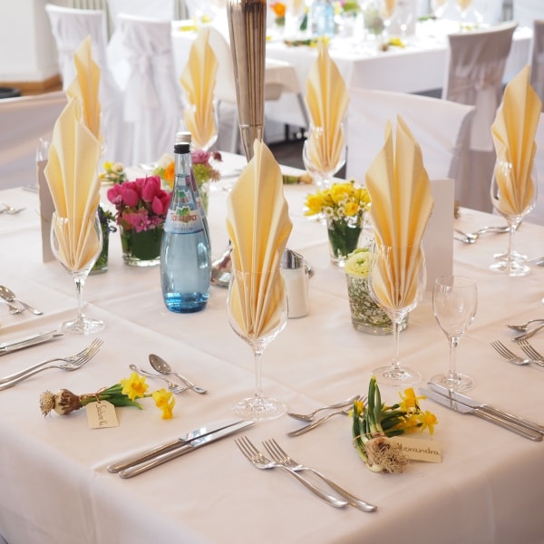 Indoor banquet table set with white linens, folded napkins in glassware, silverware, and small floral centerpieces.