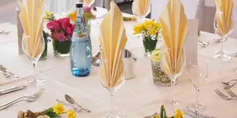 Indoor banquet table set with white linens, folded napkins in glassware, silverware, and small floral centerpieces.