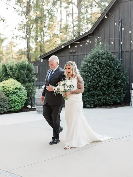 A bride and her father walking in front of a dark wooden barn with string lights.